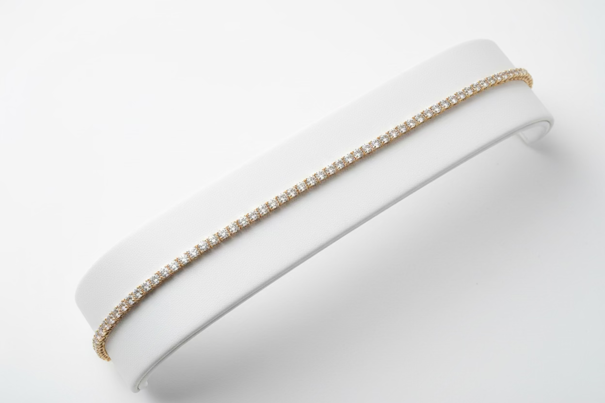 A single-strand tennis bracelet with clear rhinestones set in a gold-tone metal, displayed on a white paper band against a black background.